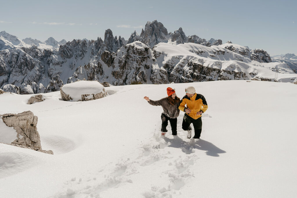 Couple walking on snowy mountain with rocky peaks.