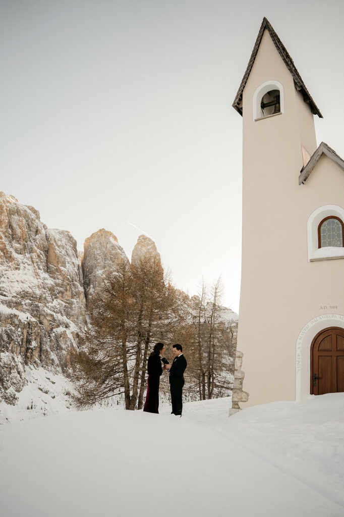 Couple stands outside snowy mountain chapel.