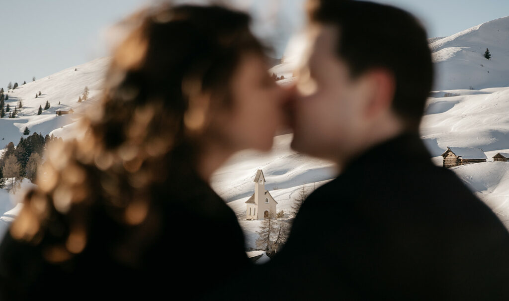 Couple kissing, snowy mountains and chapel in background.