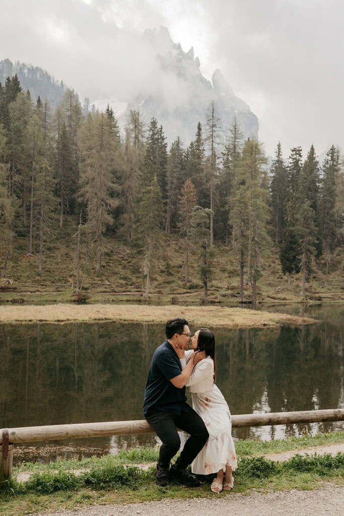 Couple kissing by lake, forest and mountain backdrop.