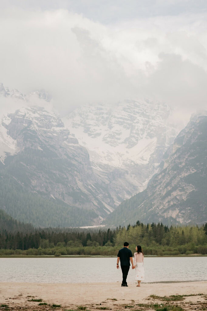 Couple walking by lake with mountain backdrop.