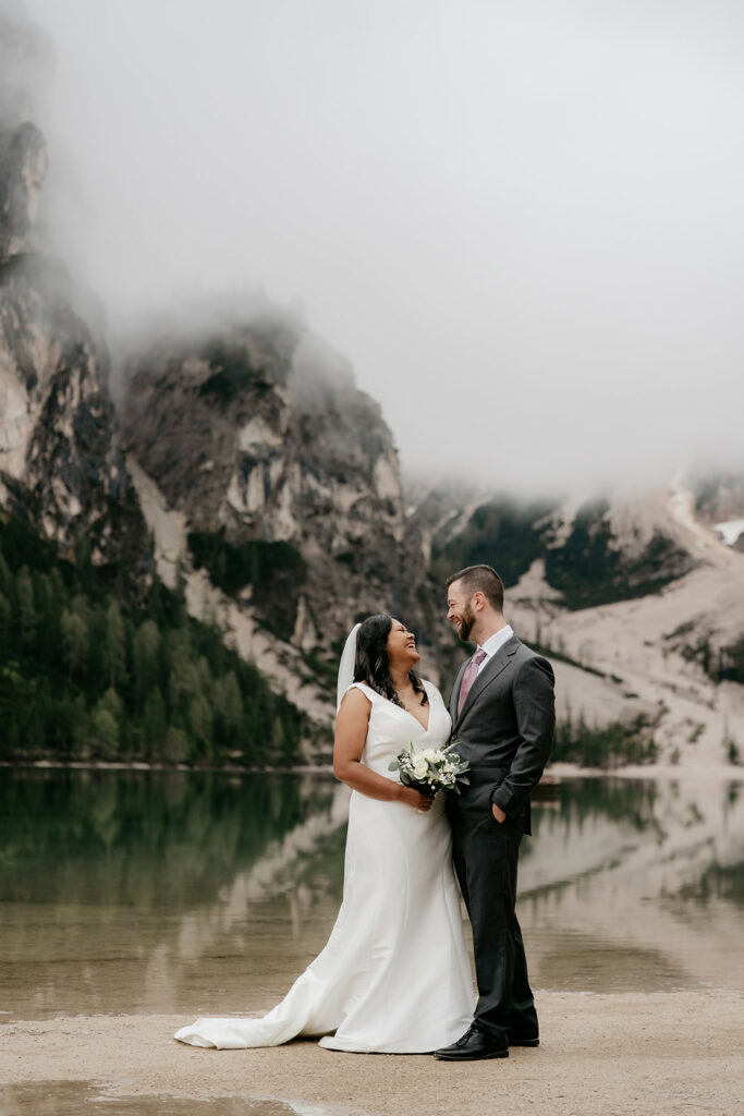 Bride and groom smiling by misty mountain lake.