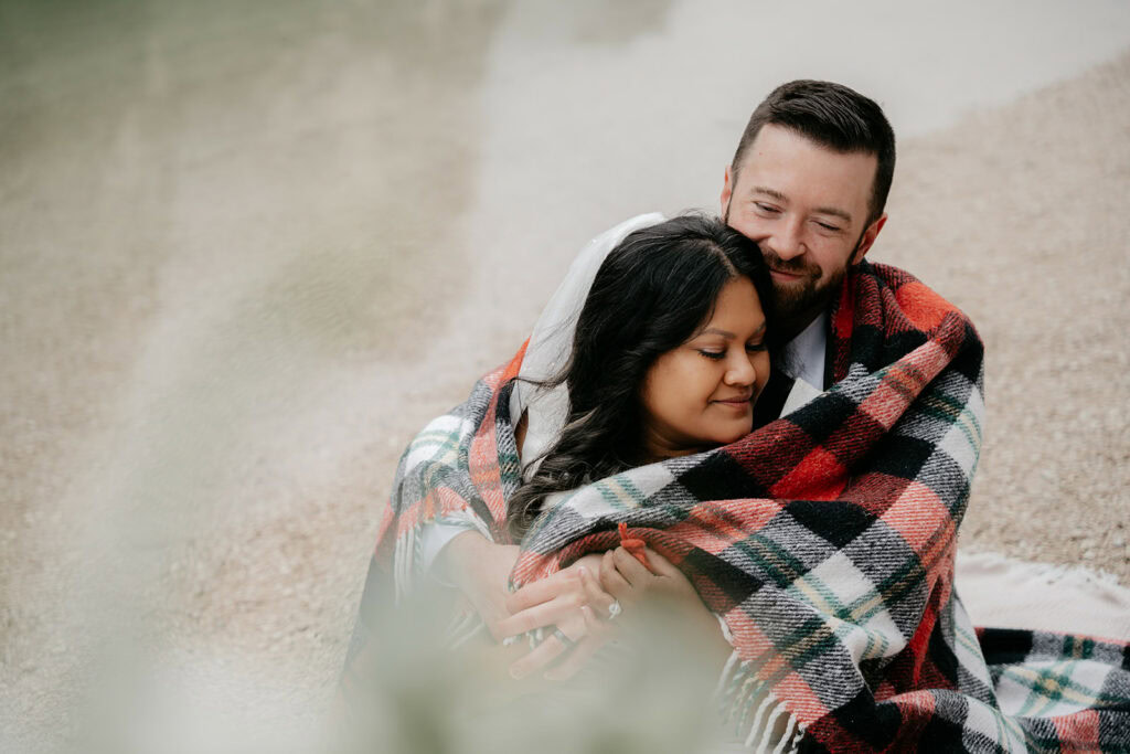 Couple wrapped in plaid blanket on beach.