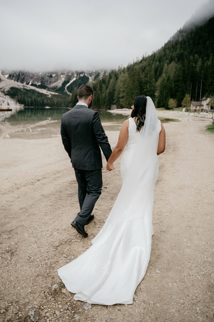 Bride and groom walk by serene mountain lake.