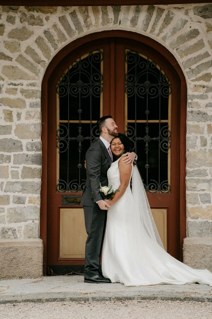 Bride and groom embracing at stone building entrance.