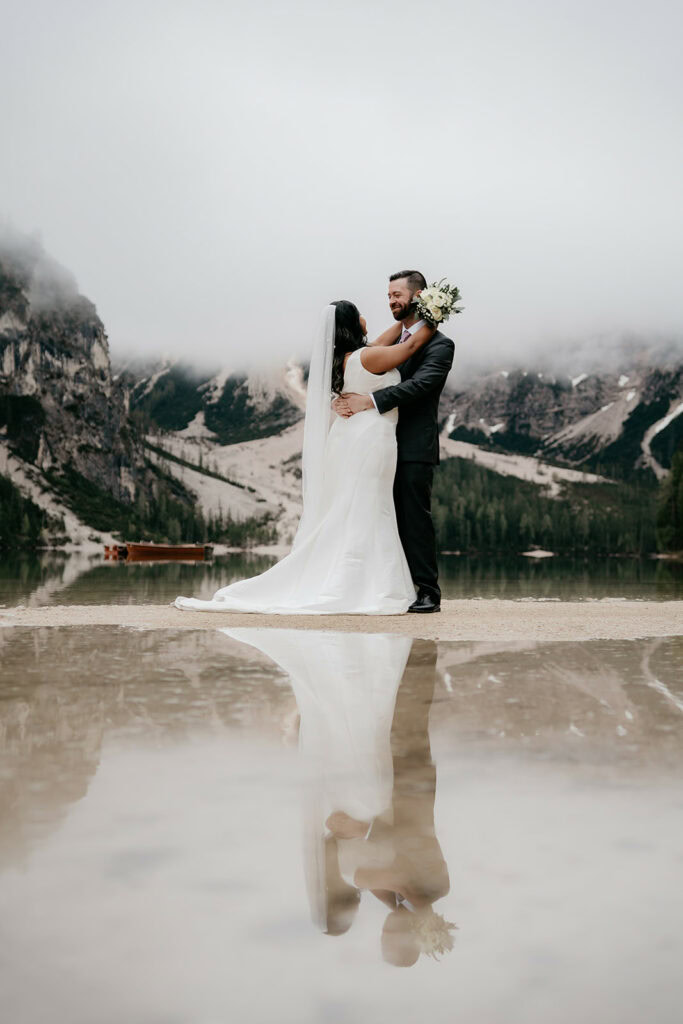 Bride and groom embrace by mountain lake.