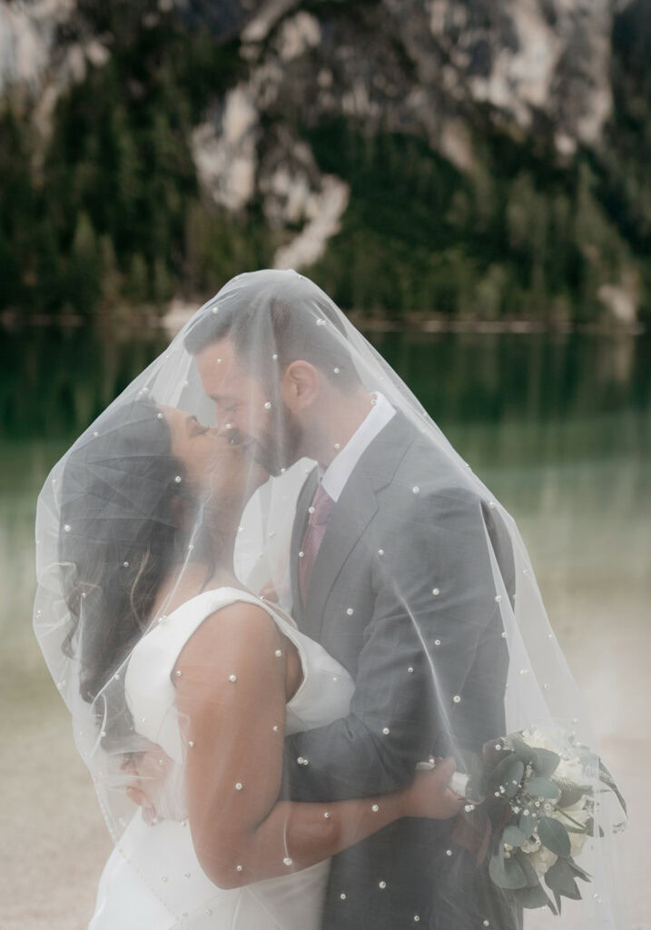 Bride and groom kissing under veil outdoors.