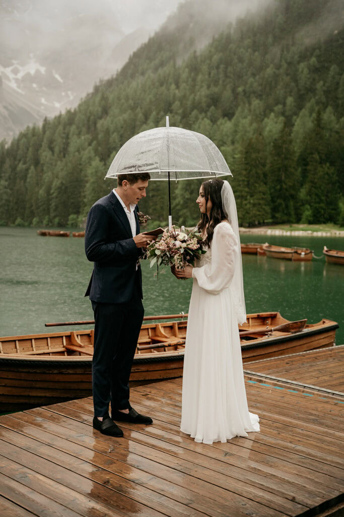 Bride and groom under umbrella on dock by lake.