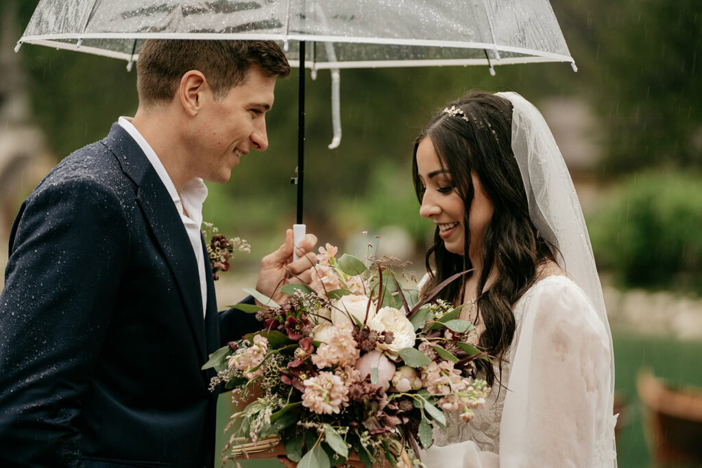 Bride and groom under umbrella, holding flowers.