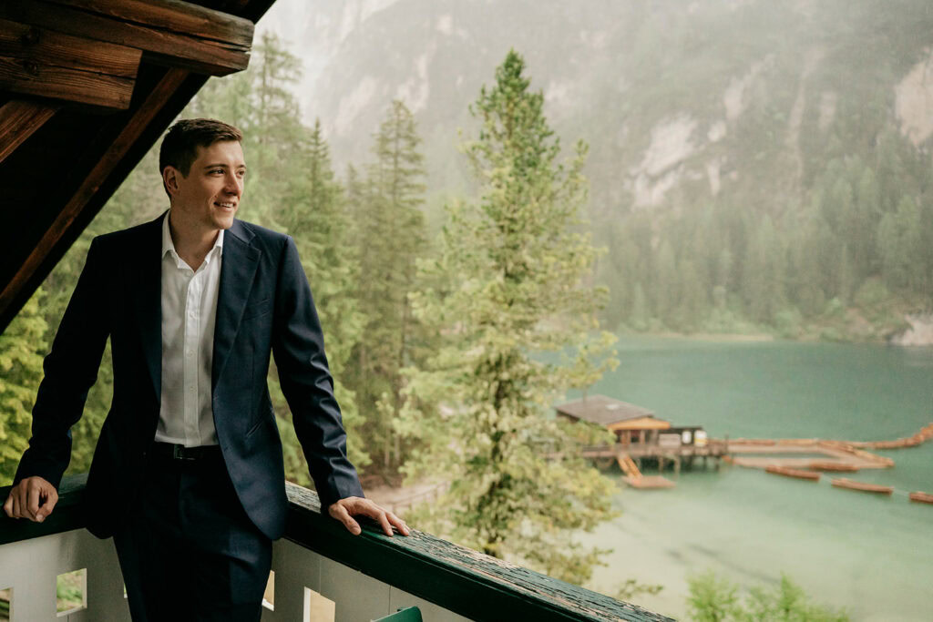 Man in suit on balcony overlooking forested lake.