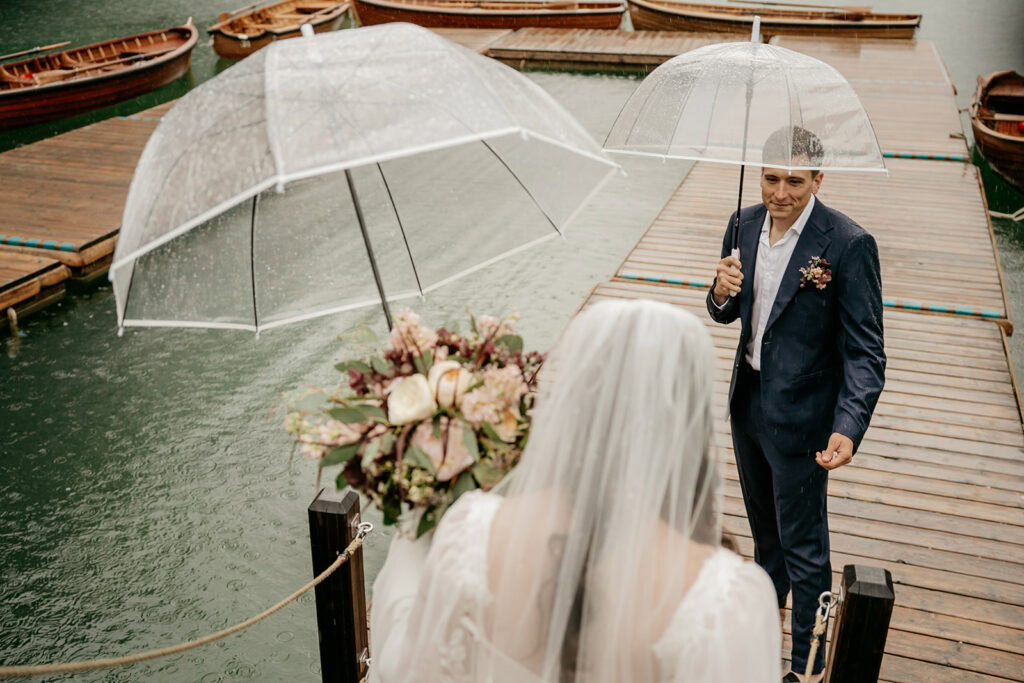 Bride and groom under umbrellas on a dock.