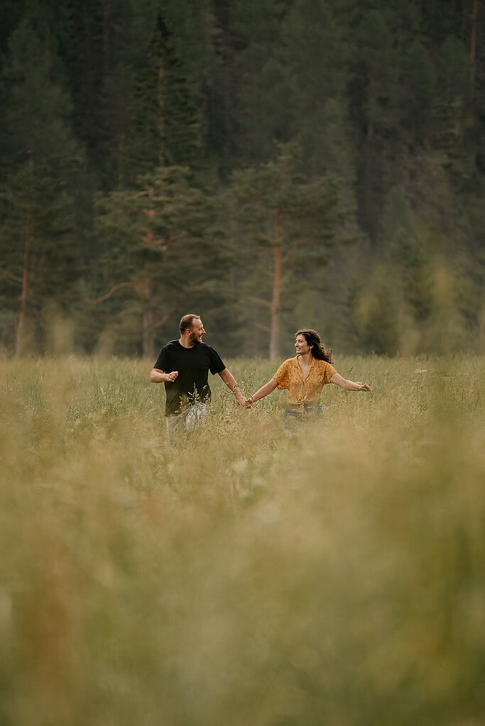 Couple holding hands in grassy field, trees behind.