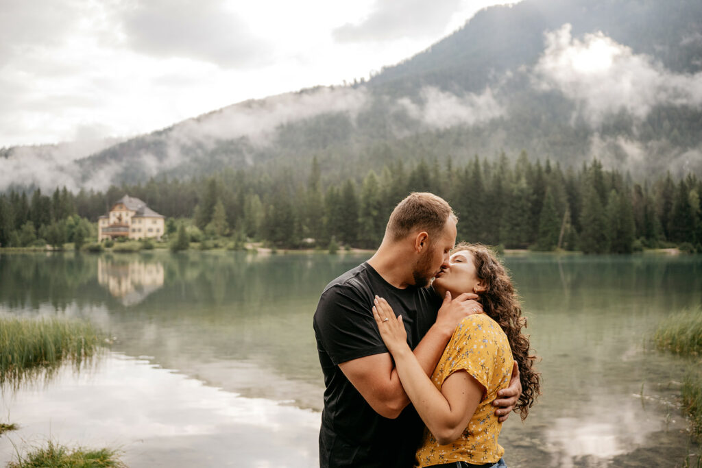 Couple kissing by a serene lake, mountain backdrop.