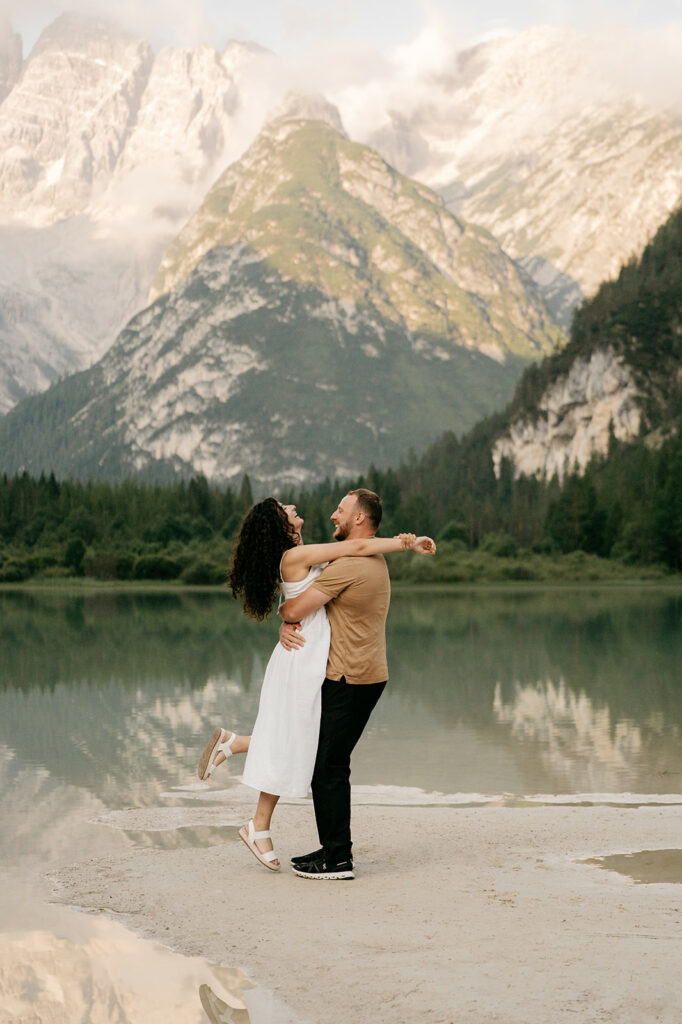 Couple embracing by lake with mountain backdrop.