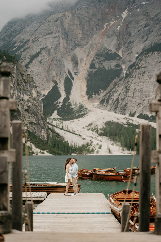 Couple kissing on lakeside dock with mountain backdrop.