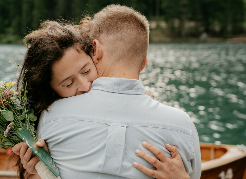 Couple hugging by a lake, holding flowers.