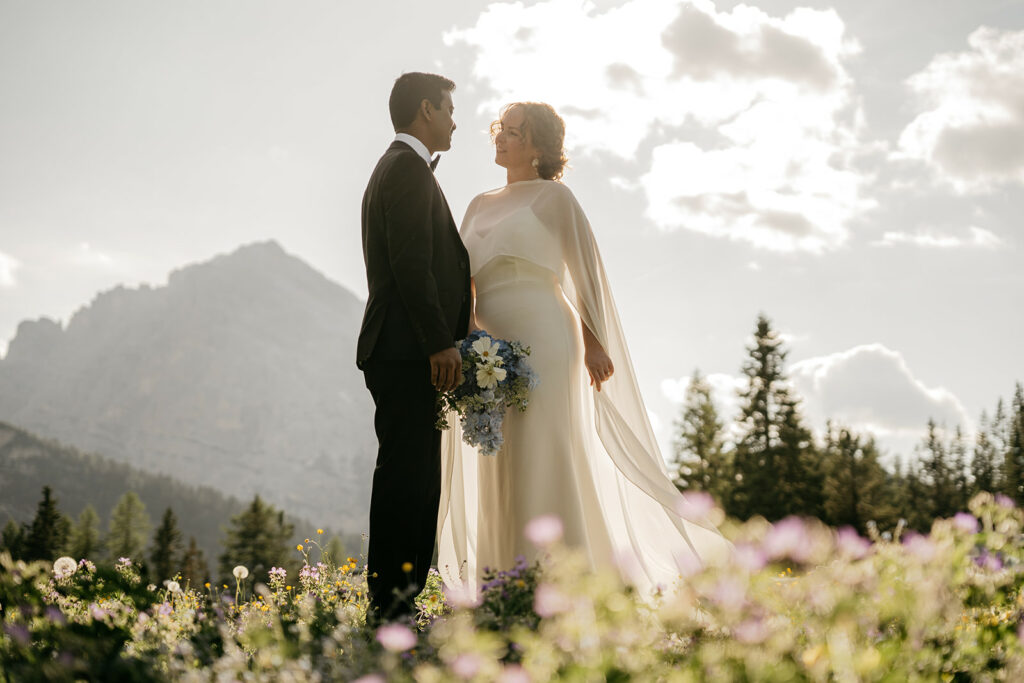 Bride and groom in mountainous field under cloudy sky.
