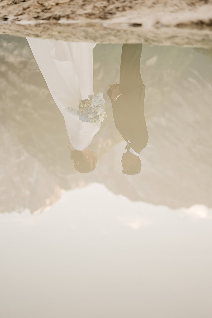 Wedding couple reflected in water, mountain backdrop.