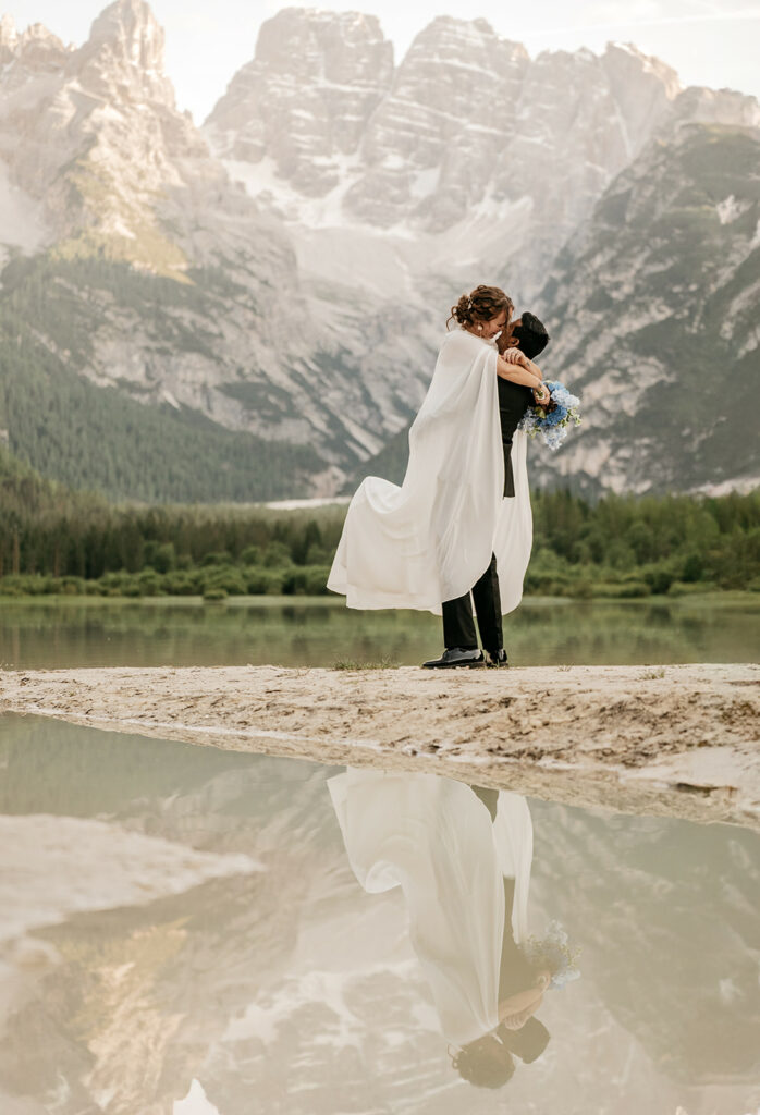 Couple embracing by mountain lake, wedding attire.