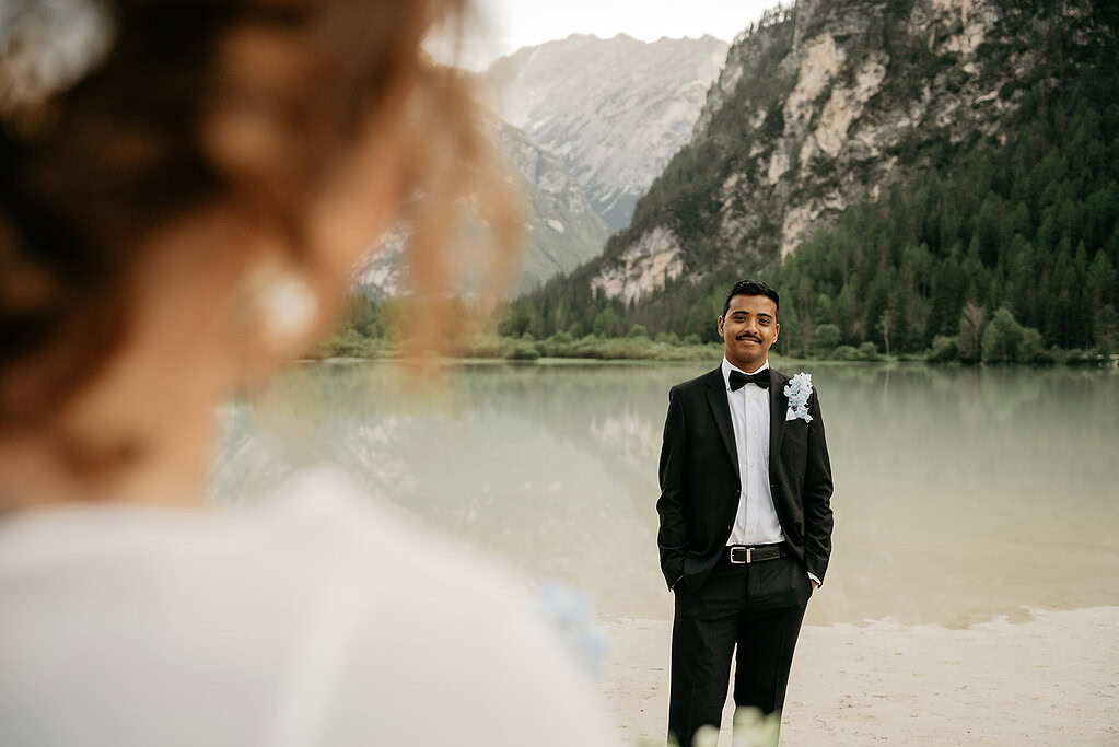 Groom in tuxedo by lake and mountains.