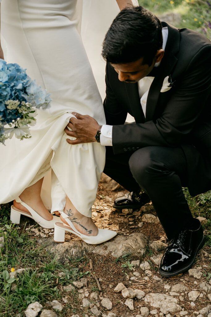 Groom adjusts bride's dress outdoors, blue bouquet nearby.