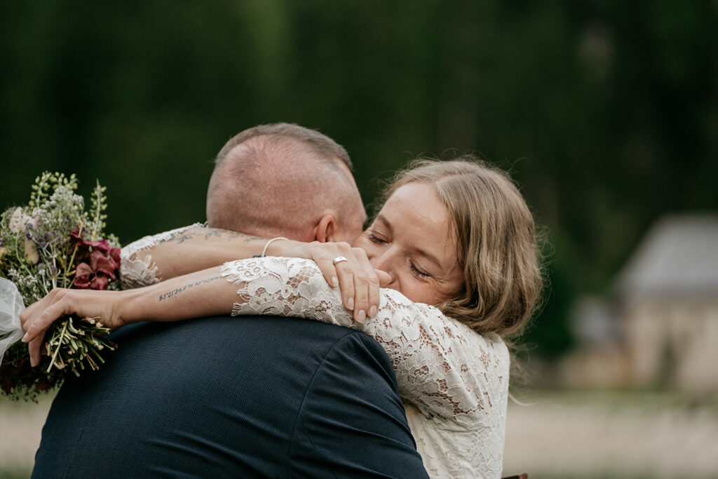 Bride hugs groom holding flowers outdoors.
