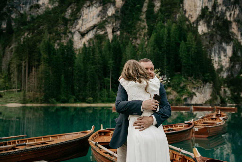 Couple embraces by scenic lake with wooden boats.