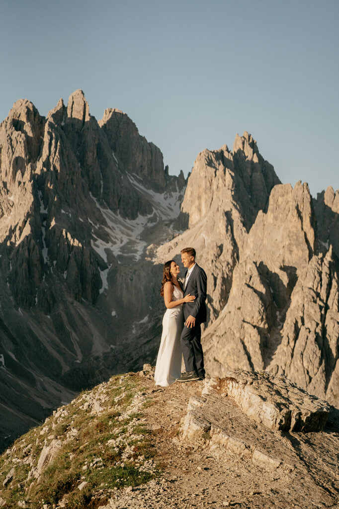 Couple standing on mountain with rocky peaks backdrop.