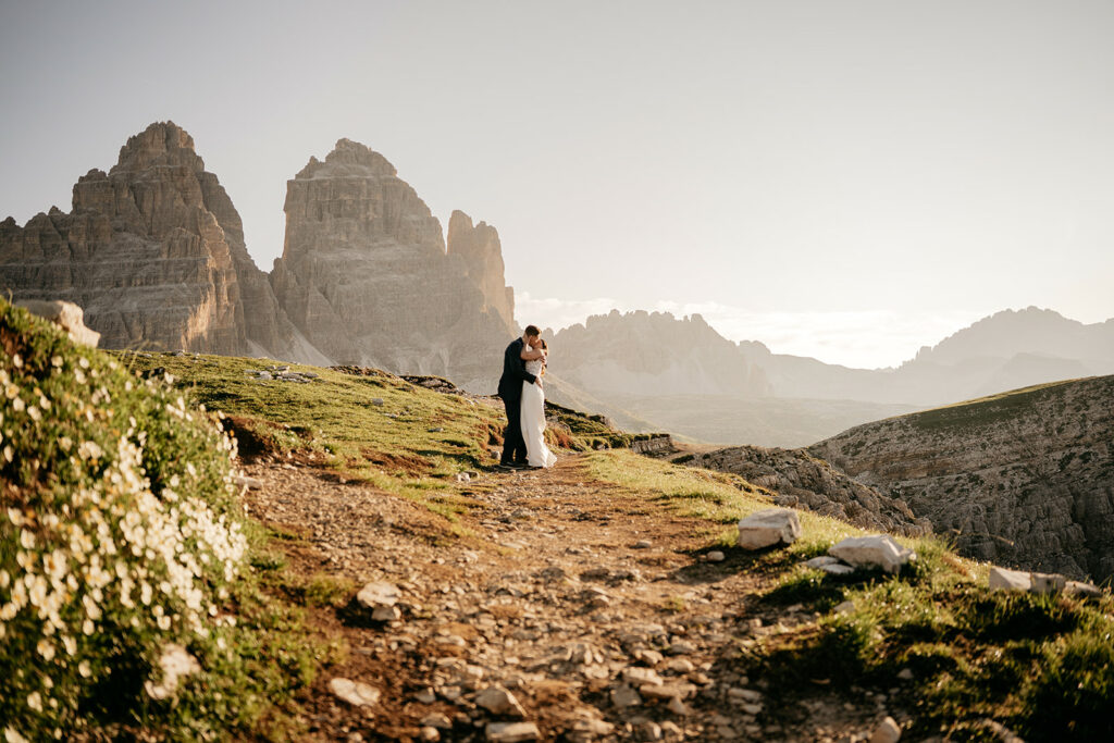Bride and groom embrace on mountain path at sunset.