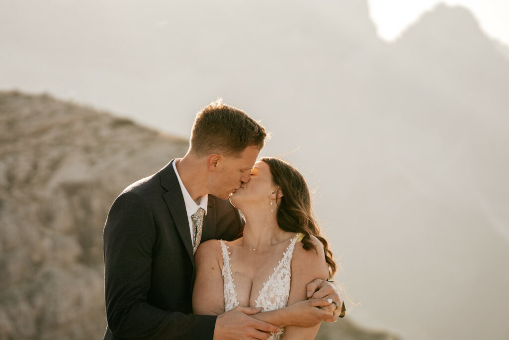 Couple kissing in wedding attire, mountain backdrop.