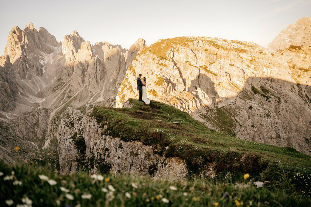 Couple embraces on cliff with mountain backdrop.