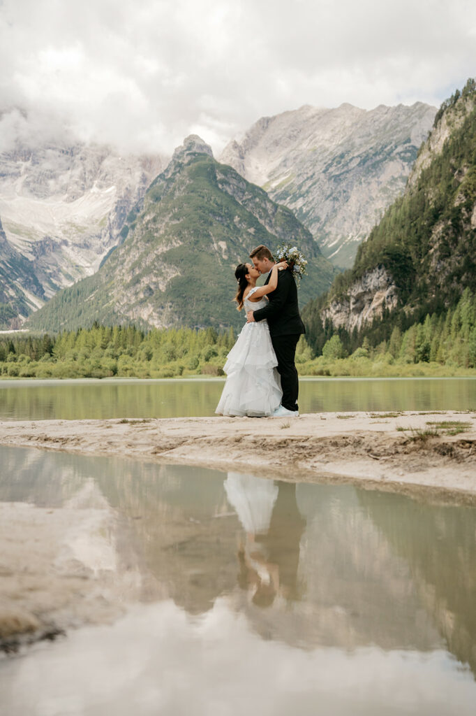 Couple embraces by mountain lake, wedding attire.
