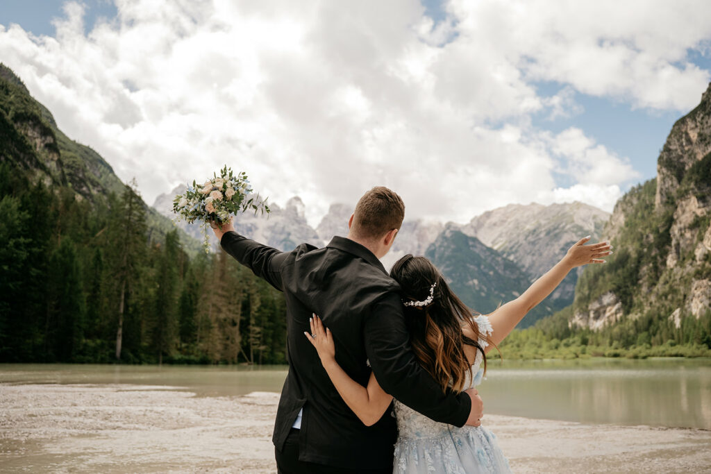 Couple embraces by lake, mountains in background.