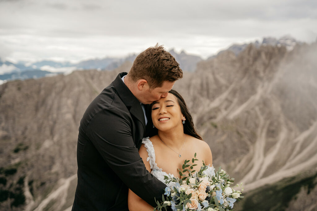 Couple embracing with mountain backdrop, bride holding bouquet.