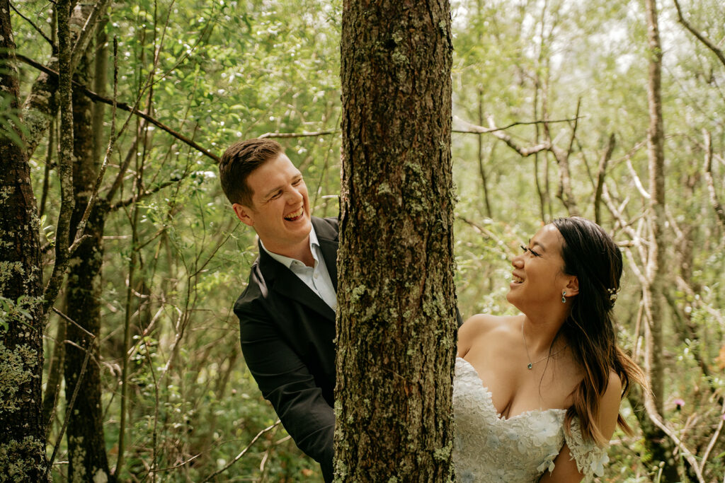 Bride and groom laughing behind a tree in forest.