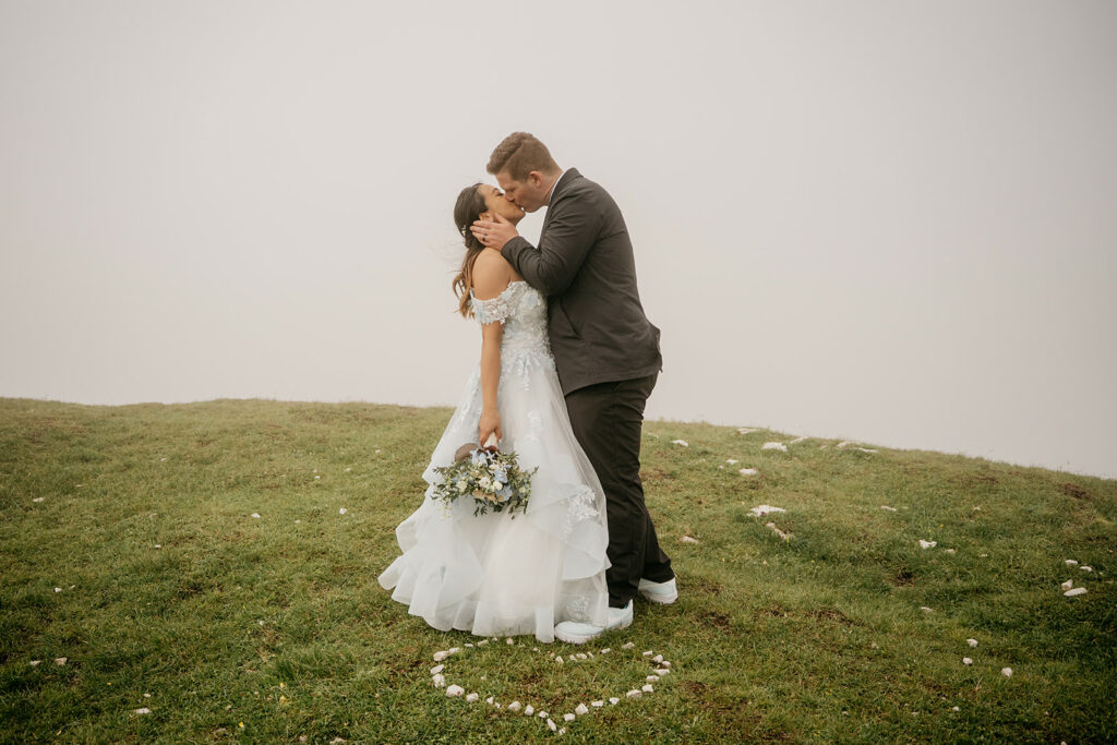 Bride and groom kissing on grassy hilltop.