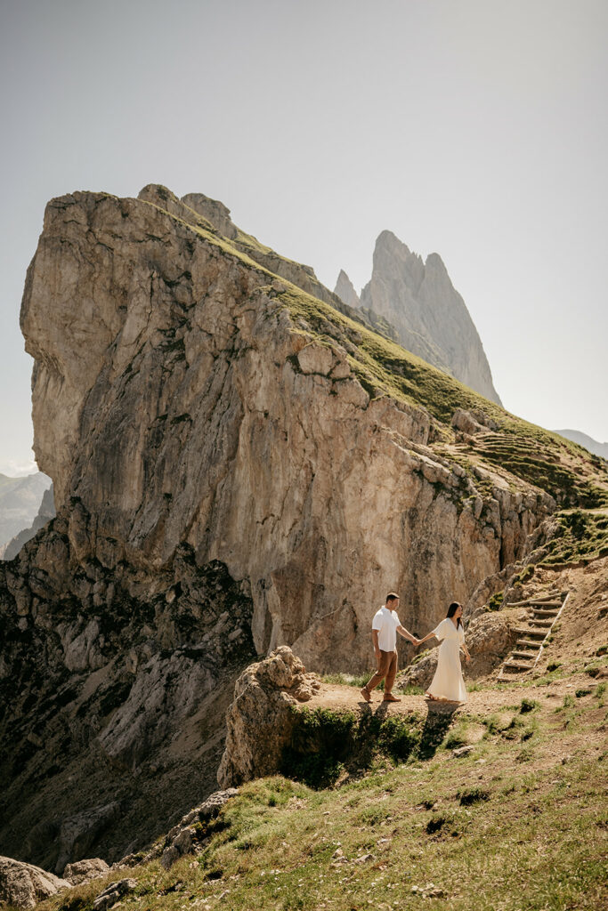 Couple walking on mountain trail with rocky backdrop.