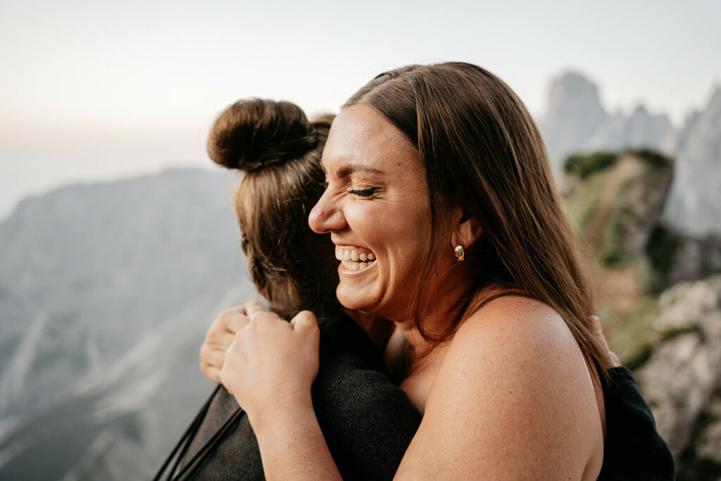 Two women hugging joyfully on a mountain path.