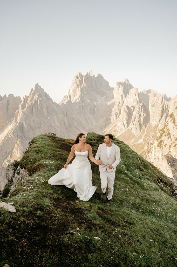Couple walks on grassy mountain, wearing wedding attire.