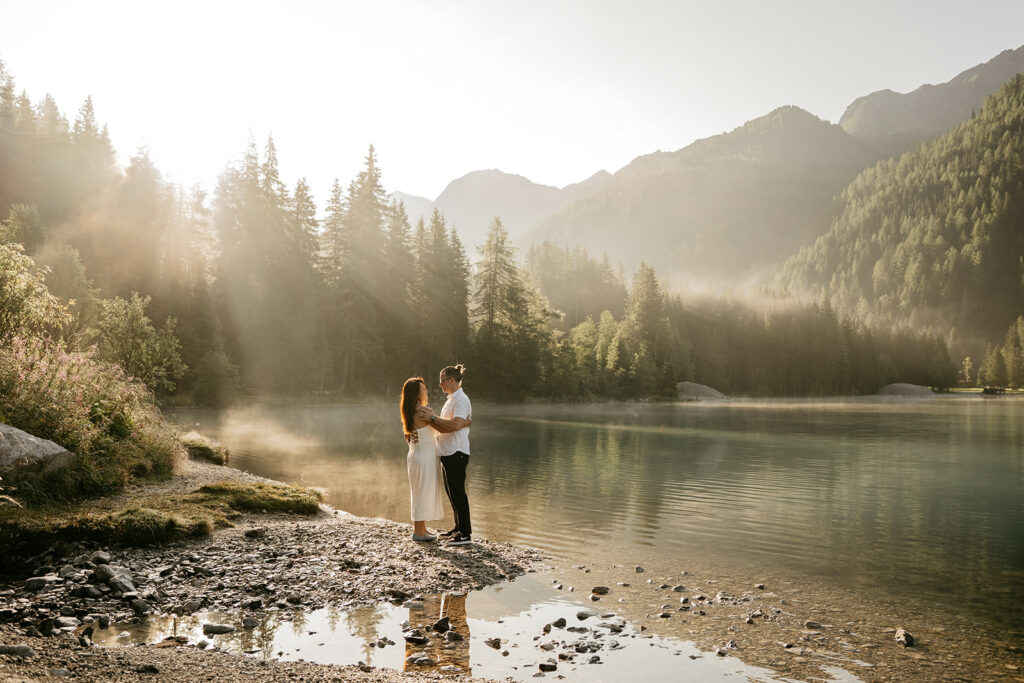 Couple embraces by misty lake with mountain backdrop.