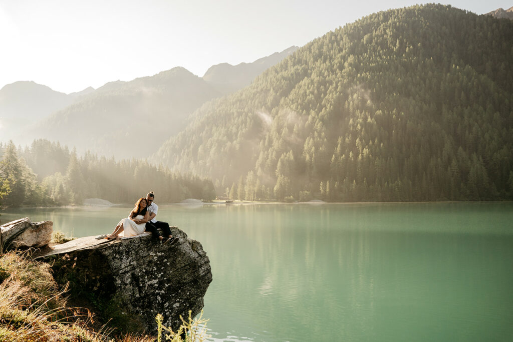 Couple embraces on rock by serene mountain lake.