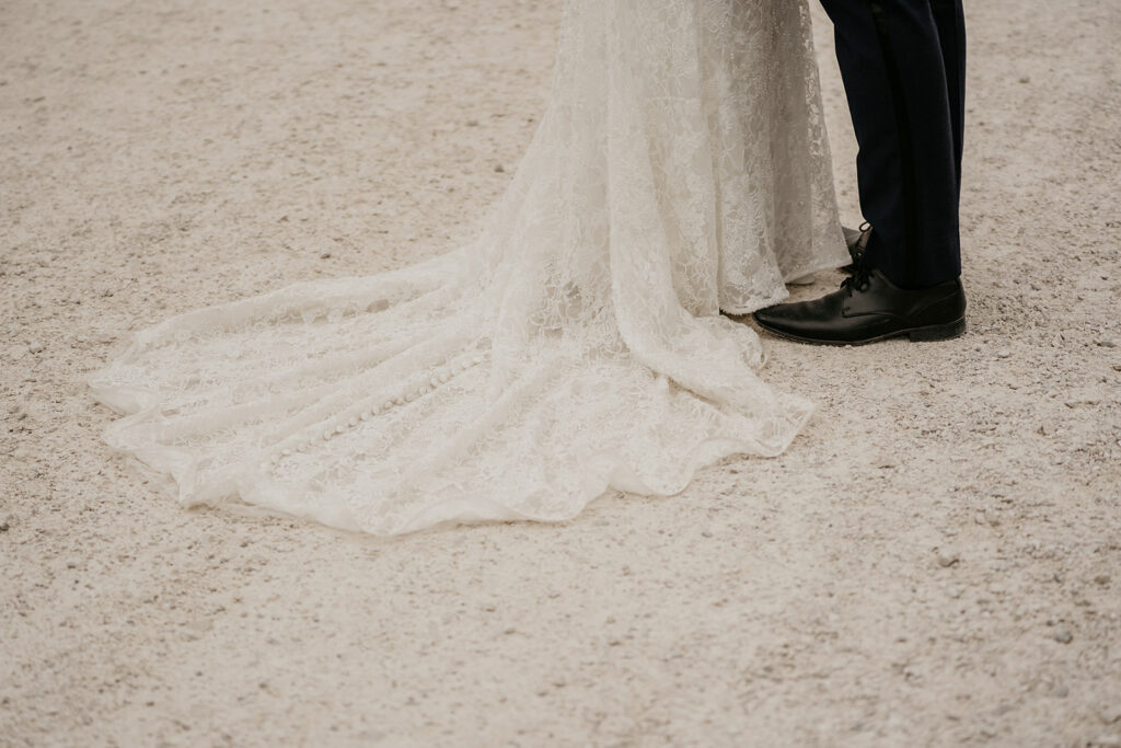 Lace wedding dress train and groom's shoes on sand.