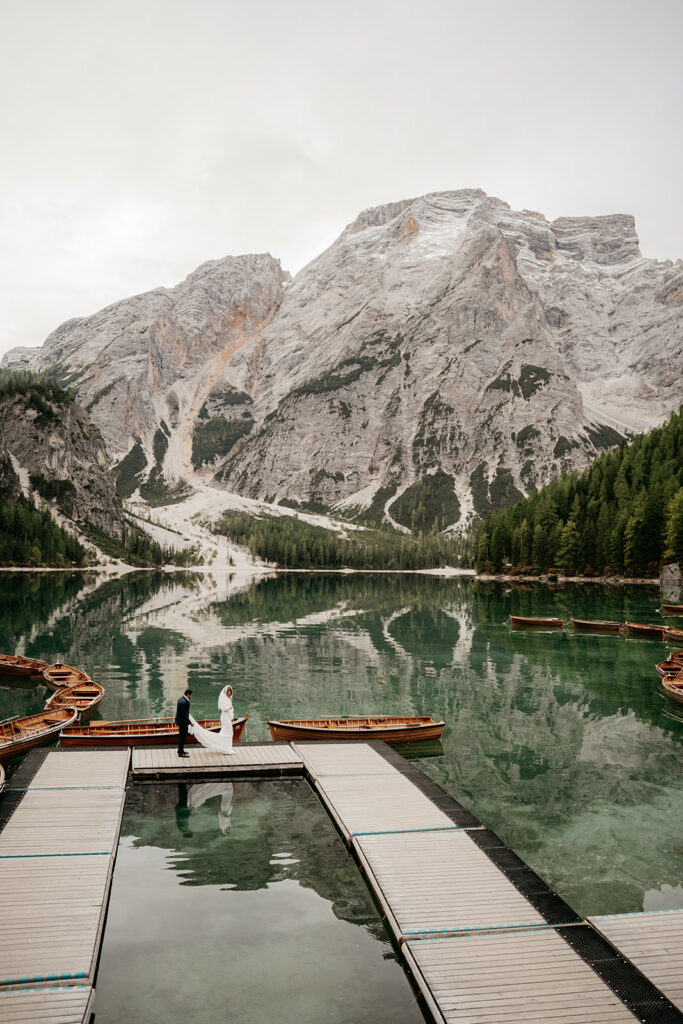 Couple on dock by mountain lake, boat reflection.