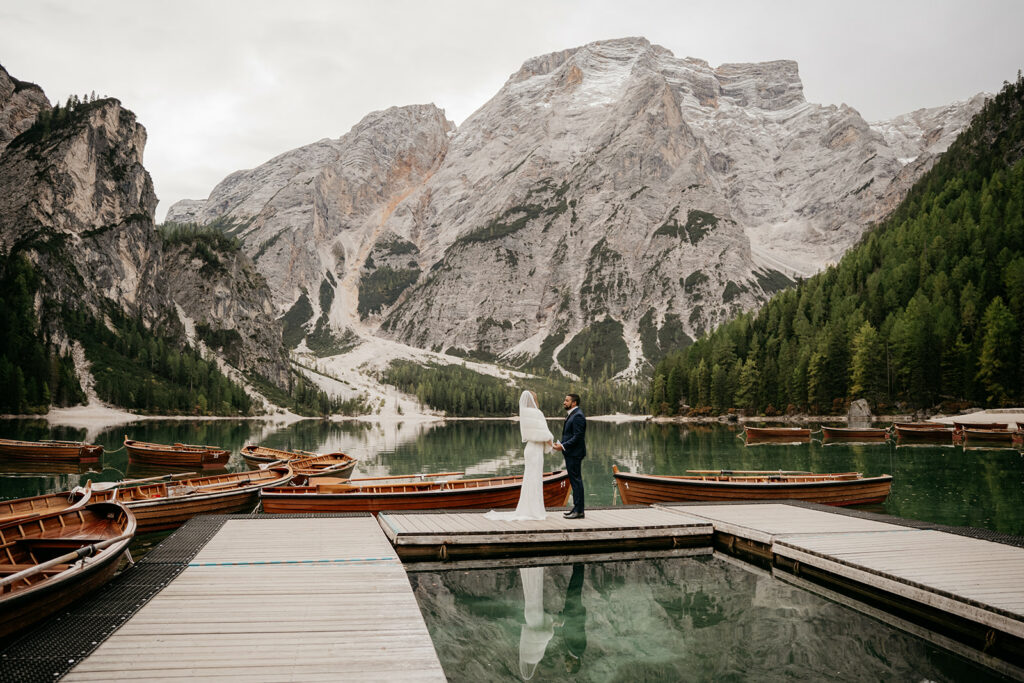 Couple on dock by mountain lake, surrounded by boats.