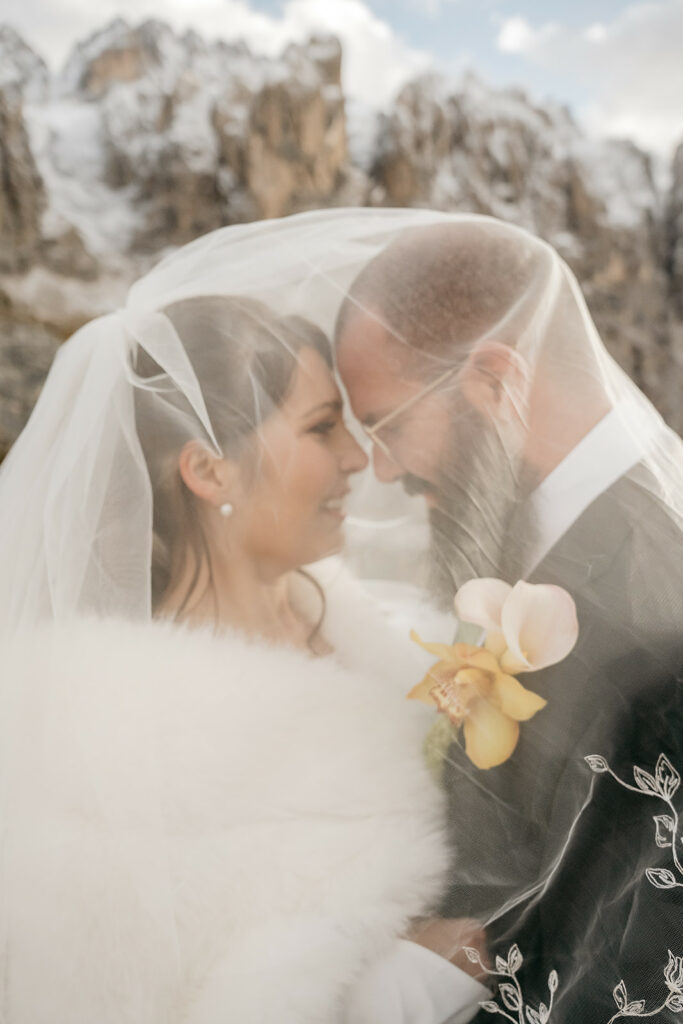 Bride and groom smiling under veil, mountain backdrop.