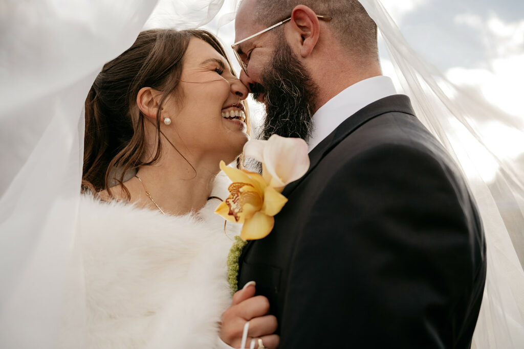 Laughing bride and groom under veil on wedding day.