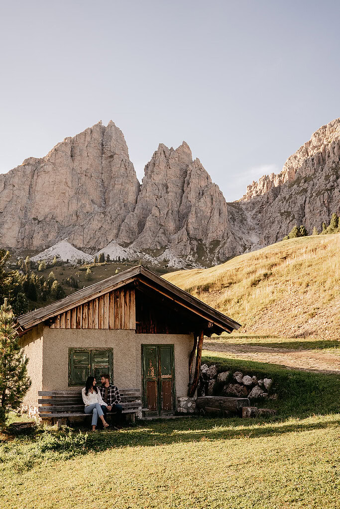 Couple sitting by rustic cabin in mountainous landscape.
