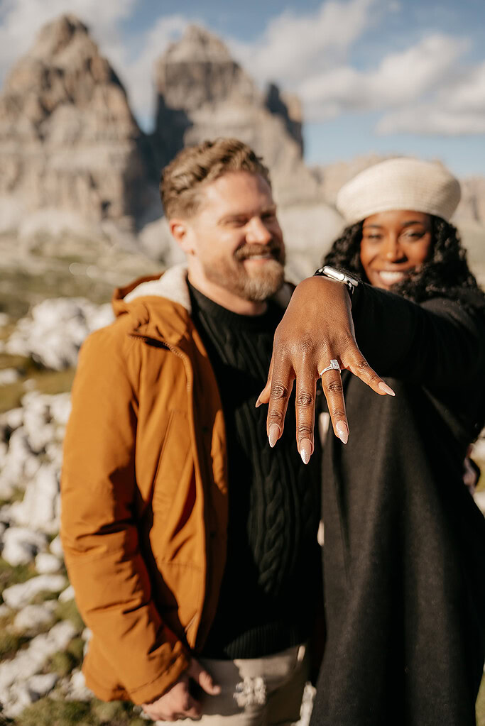 Smiling couple with engagement ring showcased in mountains.