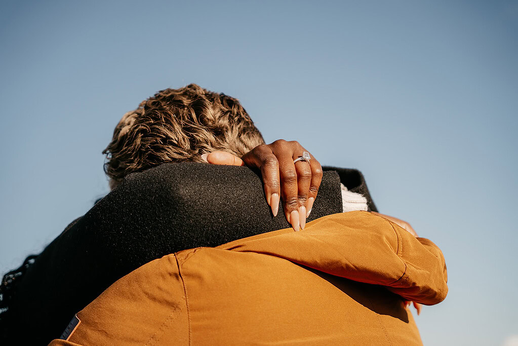 Couple embrace under blue sky, hand with ring.