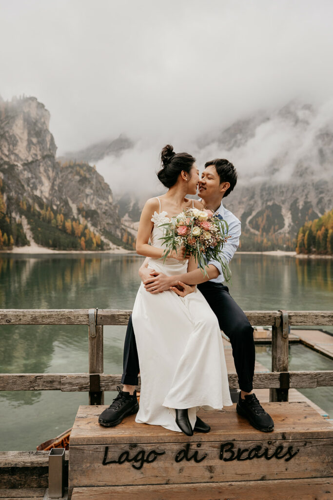 Couple embracing by mountain lake, holding bouquet.